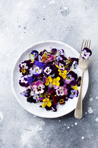 Bowl of purple, yellow, and white viola pansies with a silver fork on a grey speckled surface